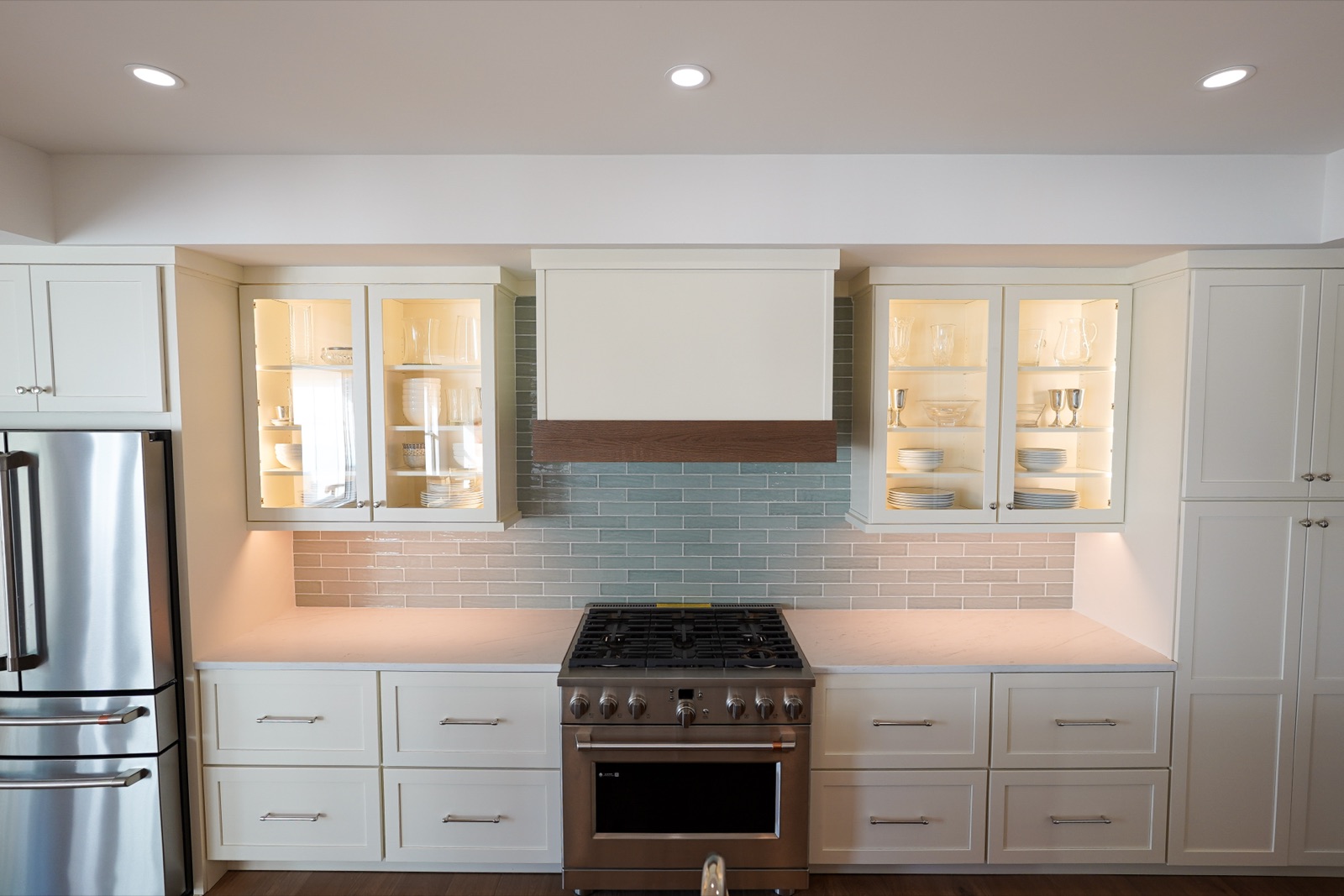 Range wall with lit glass-front uppers and a wood-trimmed painted hood by Shoreline Cabinet Company, Wilmington NC